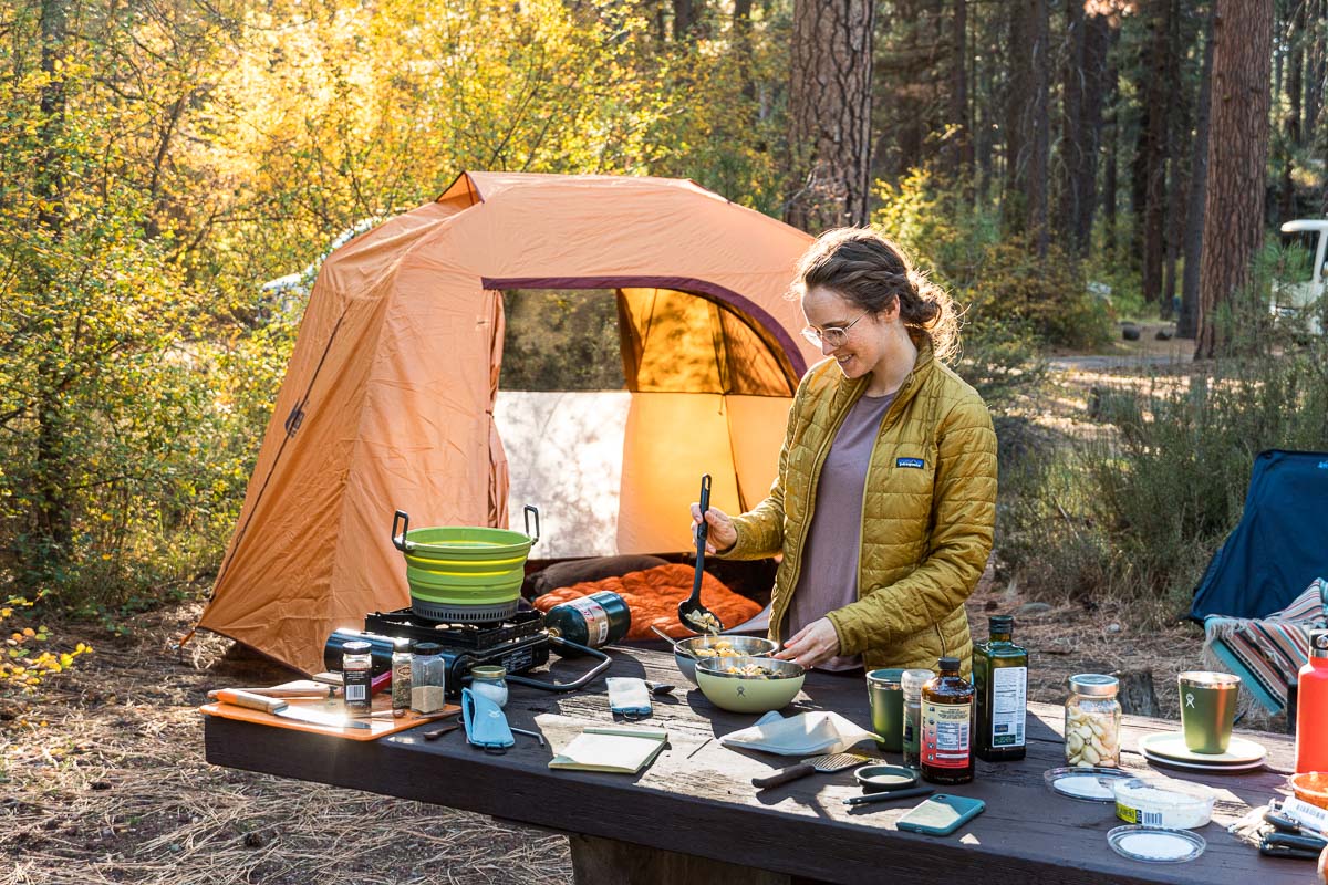Megan cooking at a picnic table. There is a camp scene and fall colors in the background