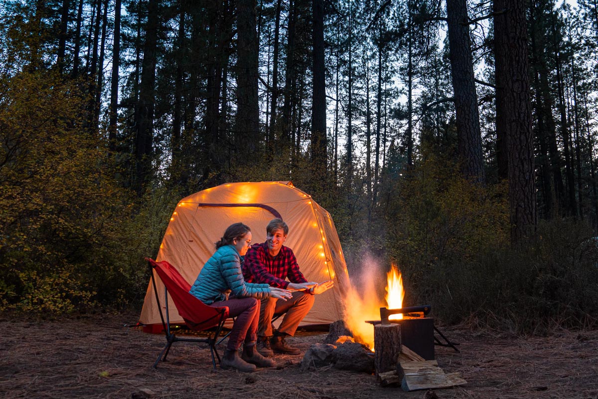 Megan and Michael sit near a campfire with a camping tent in the background