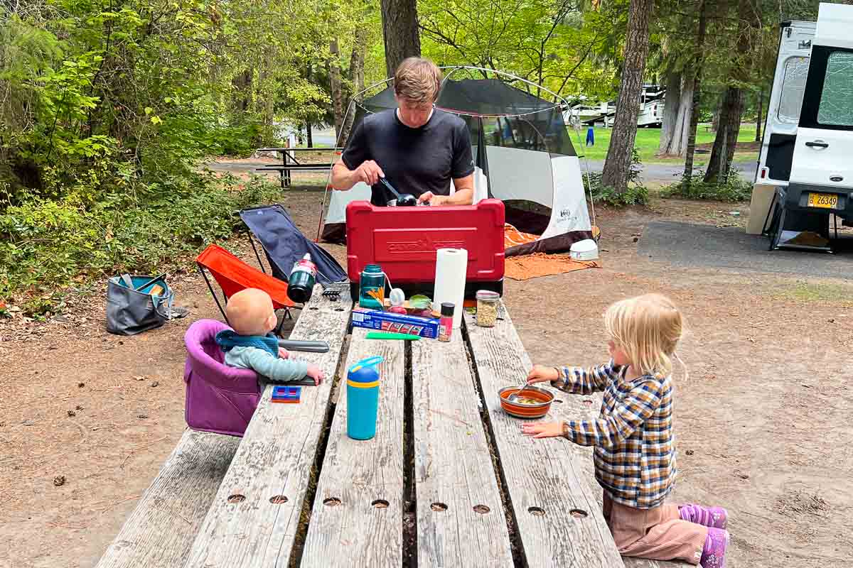 A man cooks at a red stove near a baby and young child at a wooden picnic table, with a tent, trees, and camper behind them.