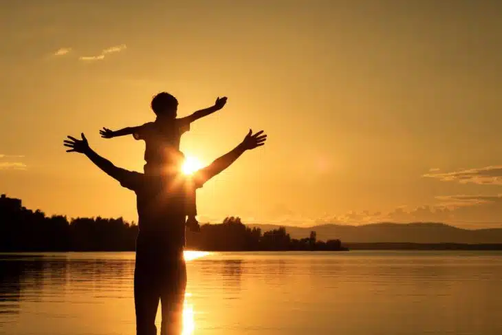 Silhouette of a child sitting on a parents shoulder with a lake in the background.