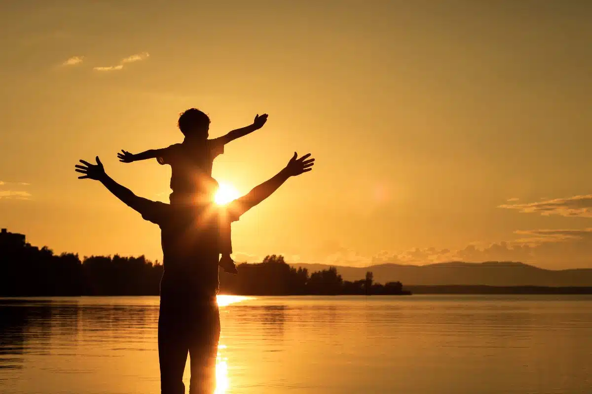 Silhouette of a child sitting on a parents shoulder with a lake in the background.