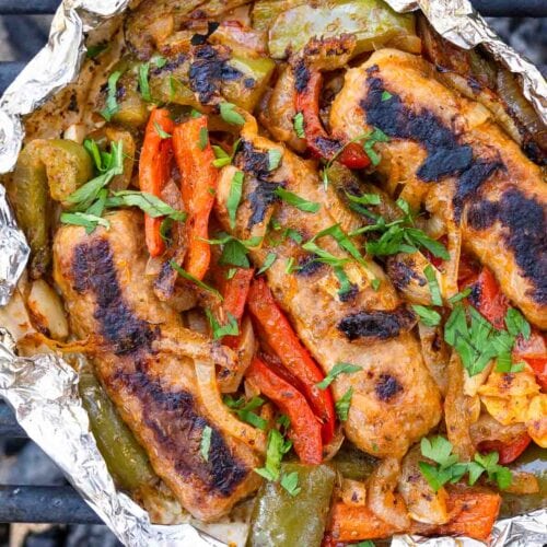 Grilled sausage, bell peppers, and onions in foil topped with chopped parsley on a grill.