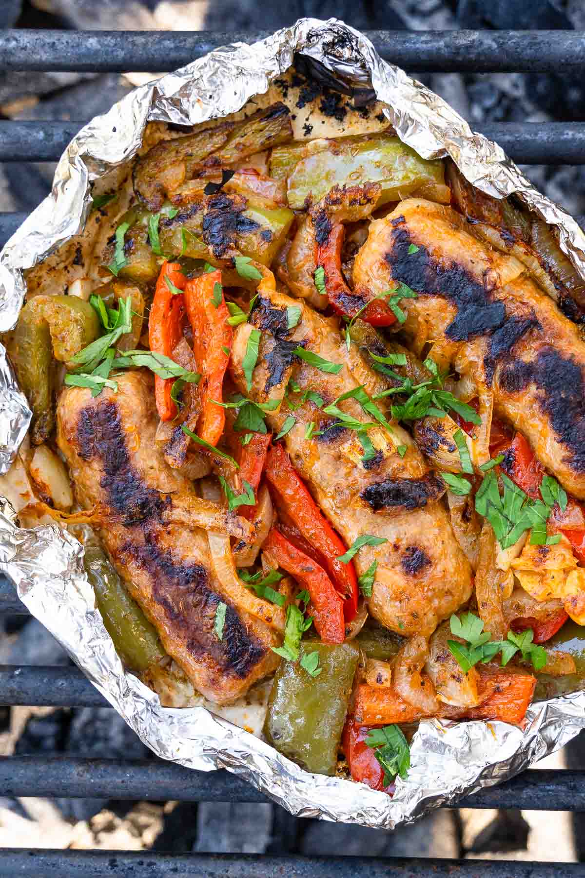 Grilled sausage, bell peppers, and onions in foil topped with chopped parsley on a grill.