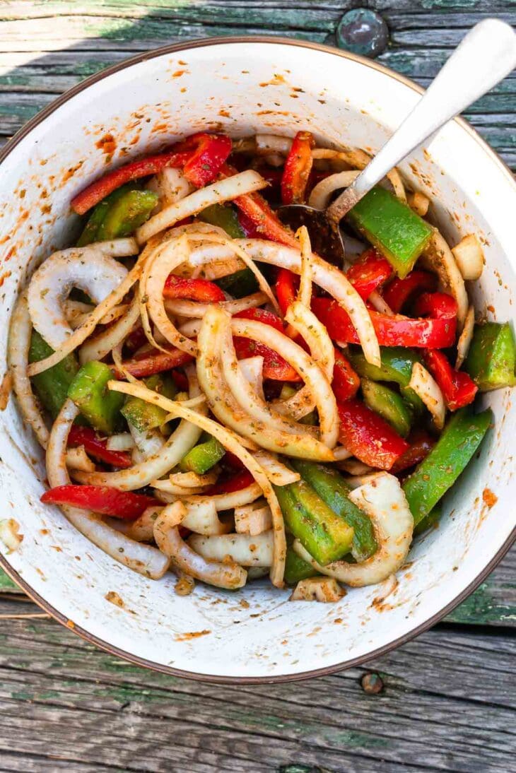 A bowl containing sliced onions, green and red bell peppers, and a spoon sits on a weathered wooden surface.