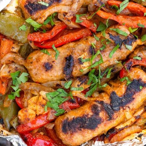 Grilled sausages with dark char marks, mixed with sautéed red and green bell peppers and onions, topped with chopped parsley in foil.