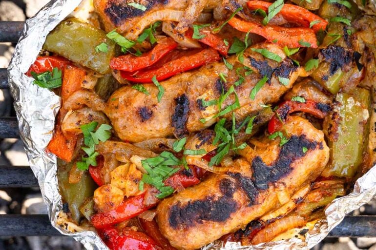 Grilled sausages with dark char marks, mixed with sautéed red and green bell peppers and onions, topped with chopped parsley in foil.