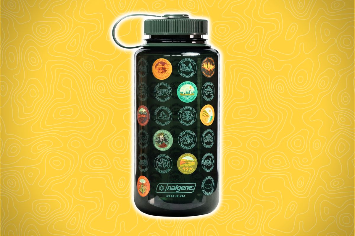A dark Nalgene bottle with a gray lid has colorful circular stickers and sits on a yellow background with a topographic pattern.