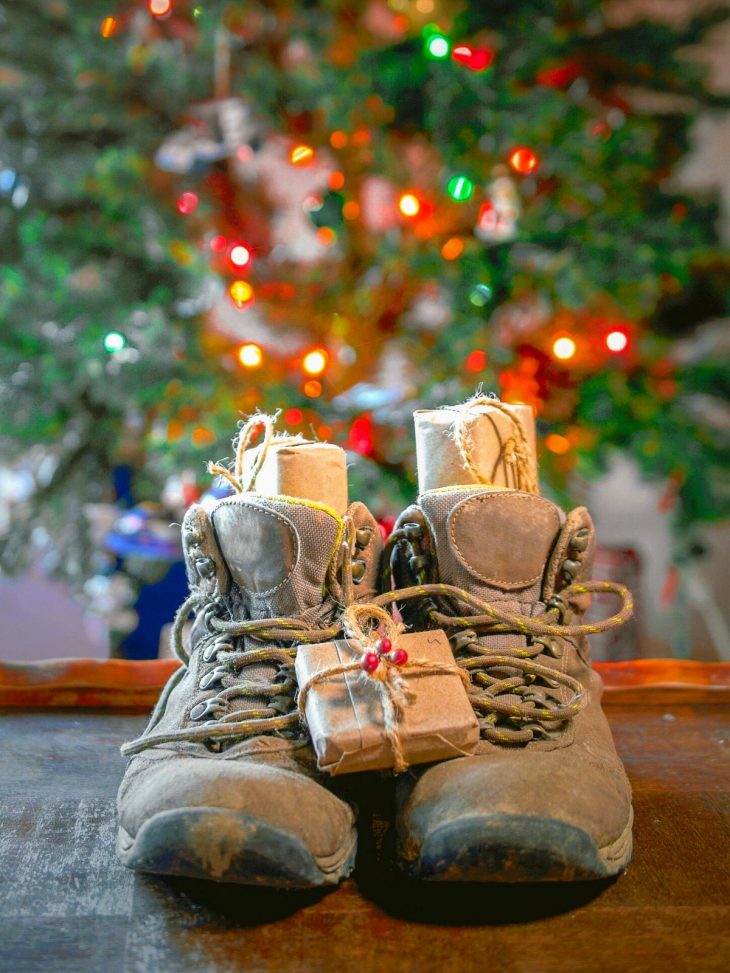 A pair of hiking boots filled with gifts. A Christmas tree is in the background