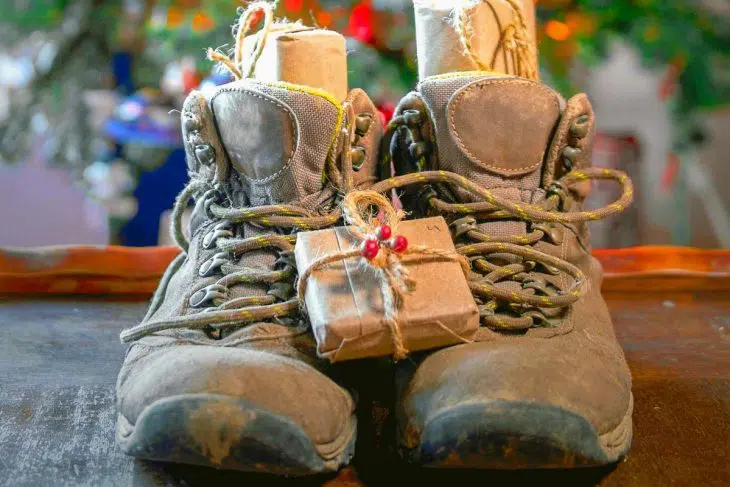 A pair of hiking boots filled with gifts. A Christmas tree is in the background