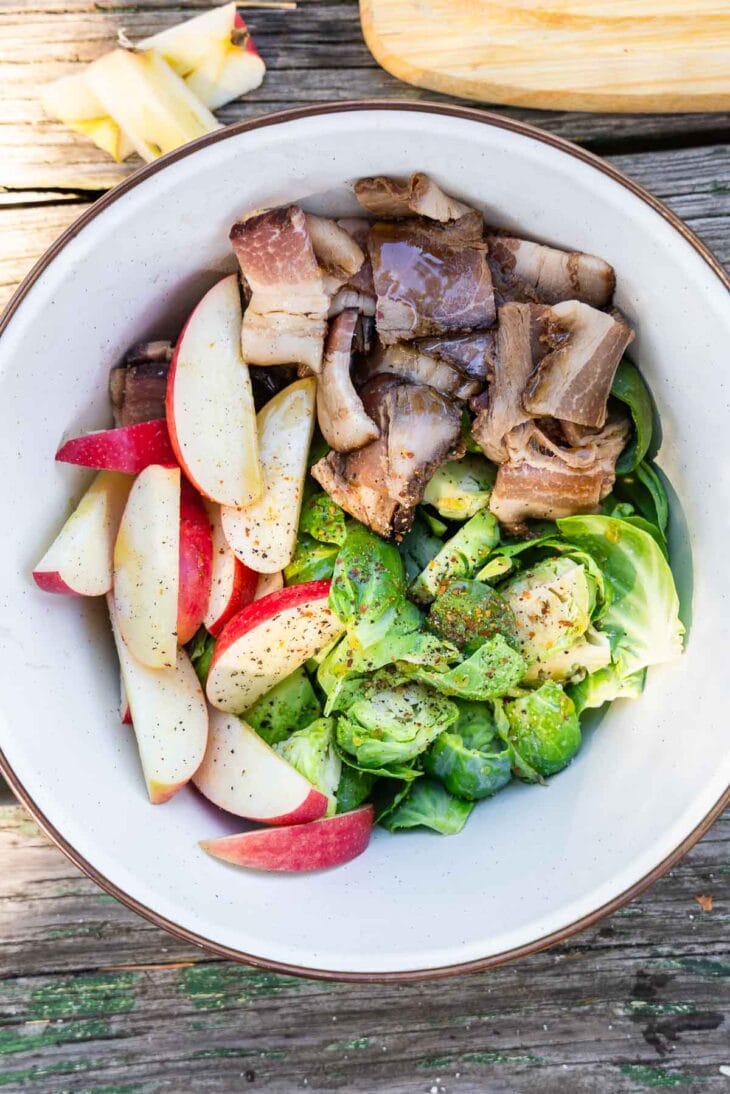 A bowl with red apple slices, mushrooms, and halved Brussels sprouts on a rustic wooden surface.