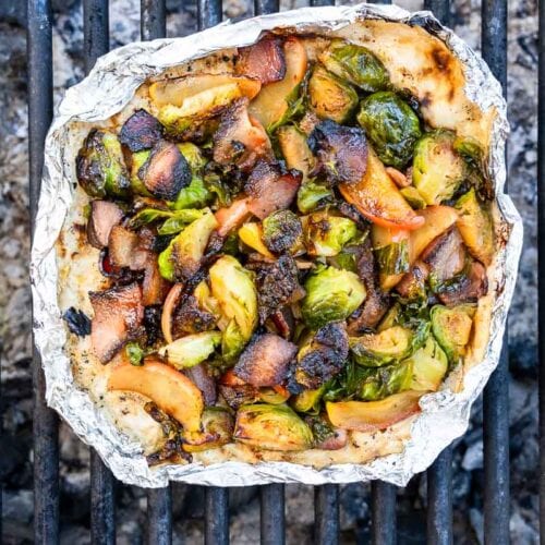 A foil packet with grilled Brussels sprouts, onions, and apple slices rests on a barbecue grill, the vegetables darkened.