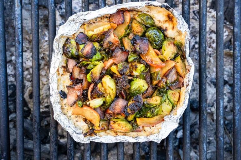 A foil packet with grilled Brussels sprouts, onions, and apple slices rests on a barbecue grill, the vegetables darkened.
