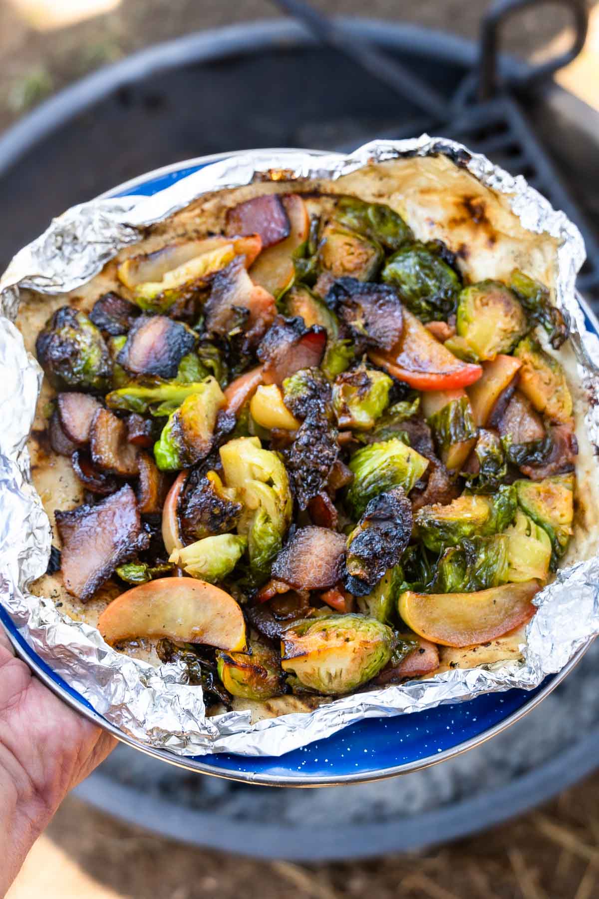 A hand holds a blue plate with a foil packet of cooked Brussels sprouts, apple slices, and bacon pieces; blurred outdoors behind.