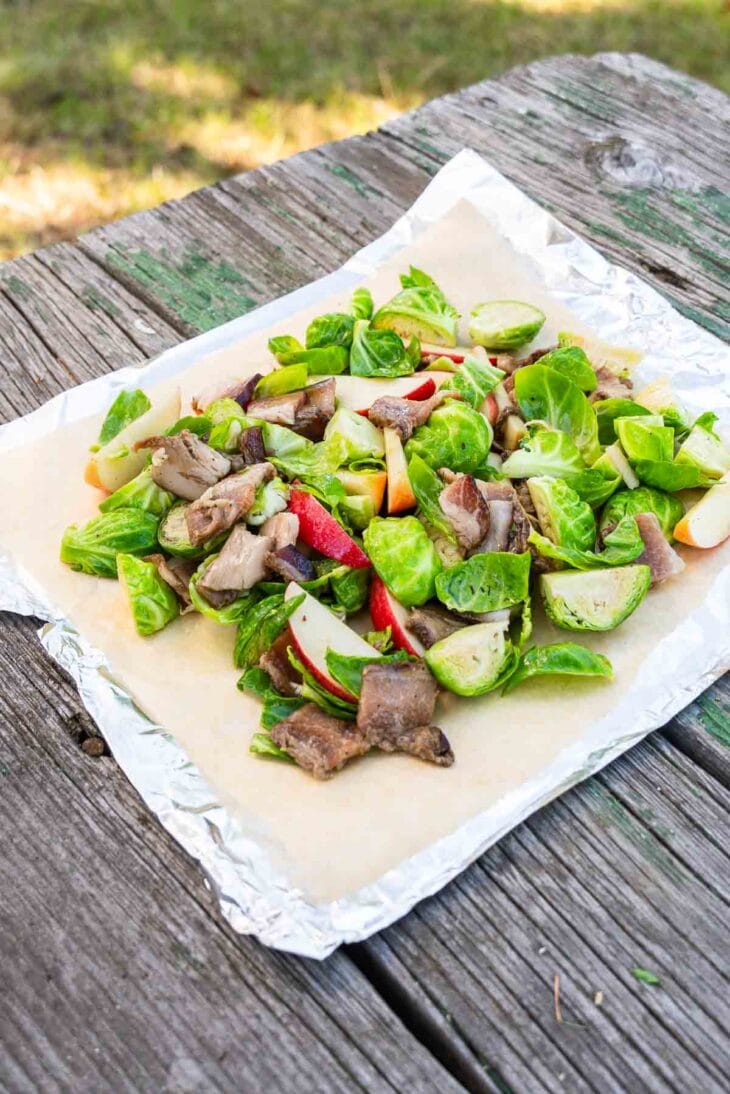 Salad with Brussels sprouts, apple slices, and mushrooms on parchment and foil atop a rustic wooden table outdoors.
