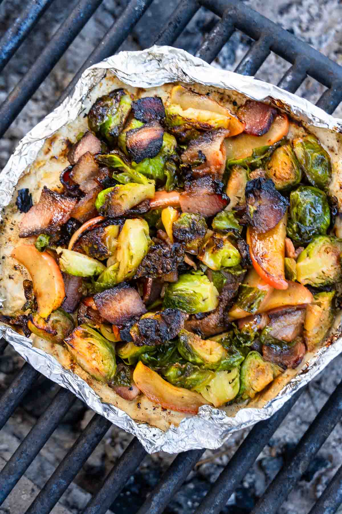 A foil packet on a grill grate contains Brussels sprouts, apple slices, and bacon pieces with charred bits and a roasted look.