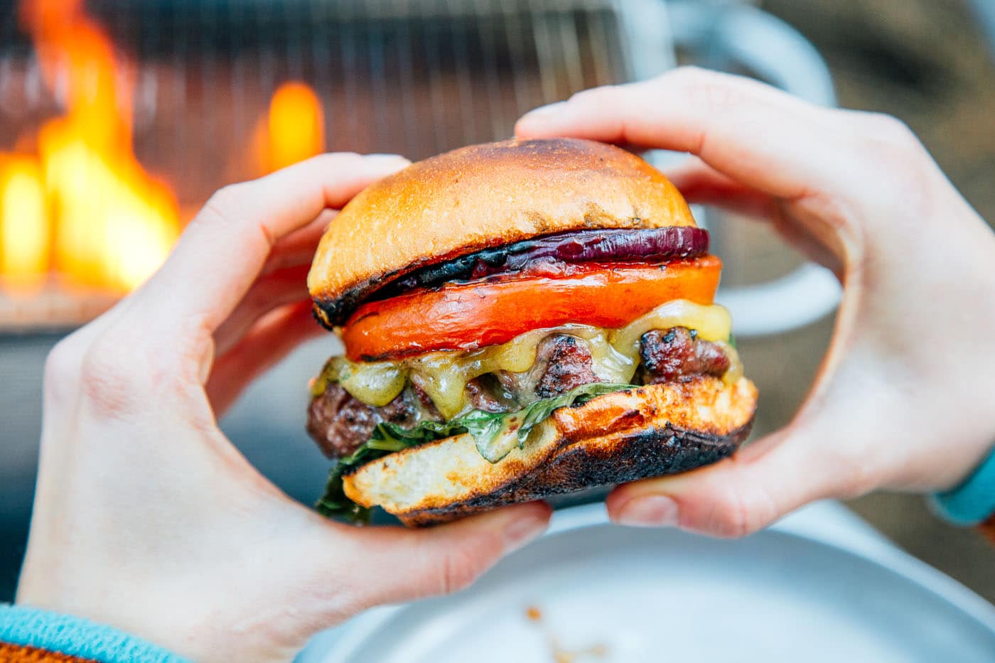 A woman holding a grilled burger in front of a campfire