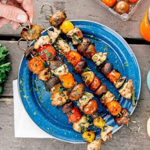 Four grilled chicken and vegetable kabobs on a blue plate. A hand is picking one up