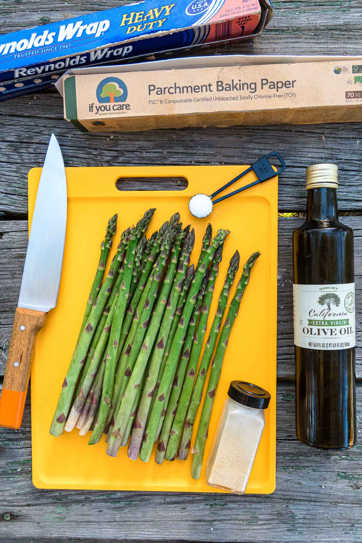 Asparagus on a cutting board next to other ingredients and boxes of parchment and foil