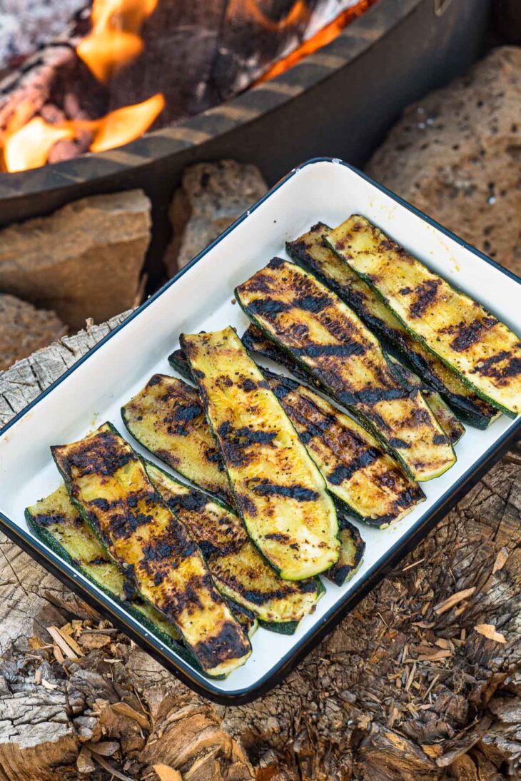 Grilled zucchini in a white enamel dish with a campfire in the background