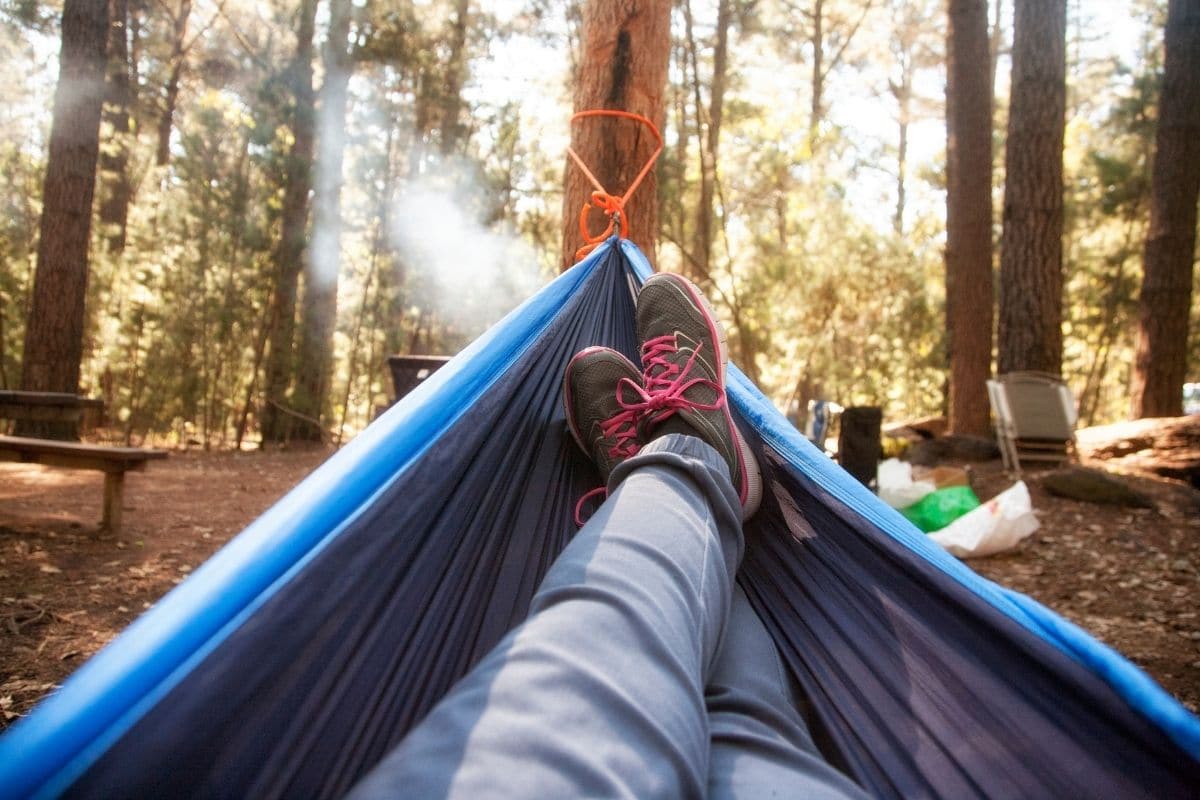 A blue hammock is strung up with a camping scene and legs can be seen in the hammock