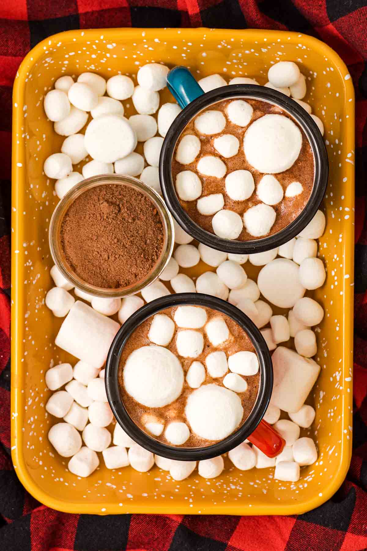 Two mugs of marshmallow-topped hot cocoa, one in a blue mug and one in a red mug, on a yellow tray with a jar of hot cocoa mix and more marshmallows.