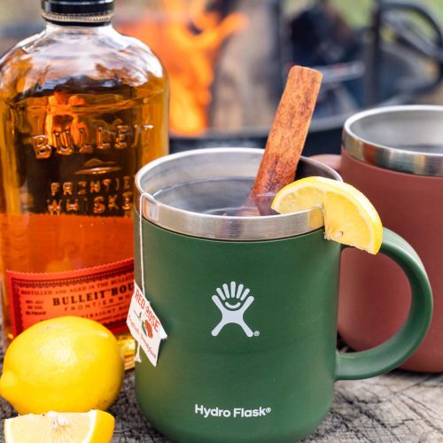 A mug of hot toddy with ingredients and a campfire in the background