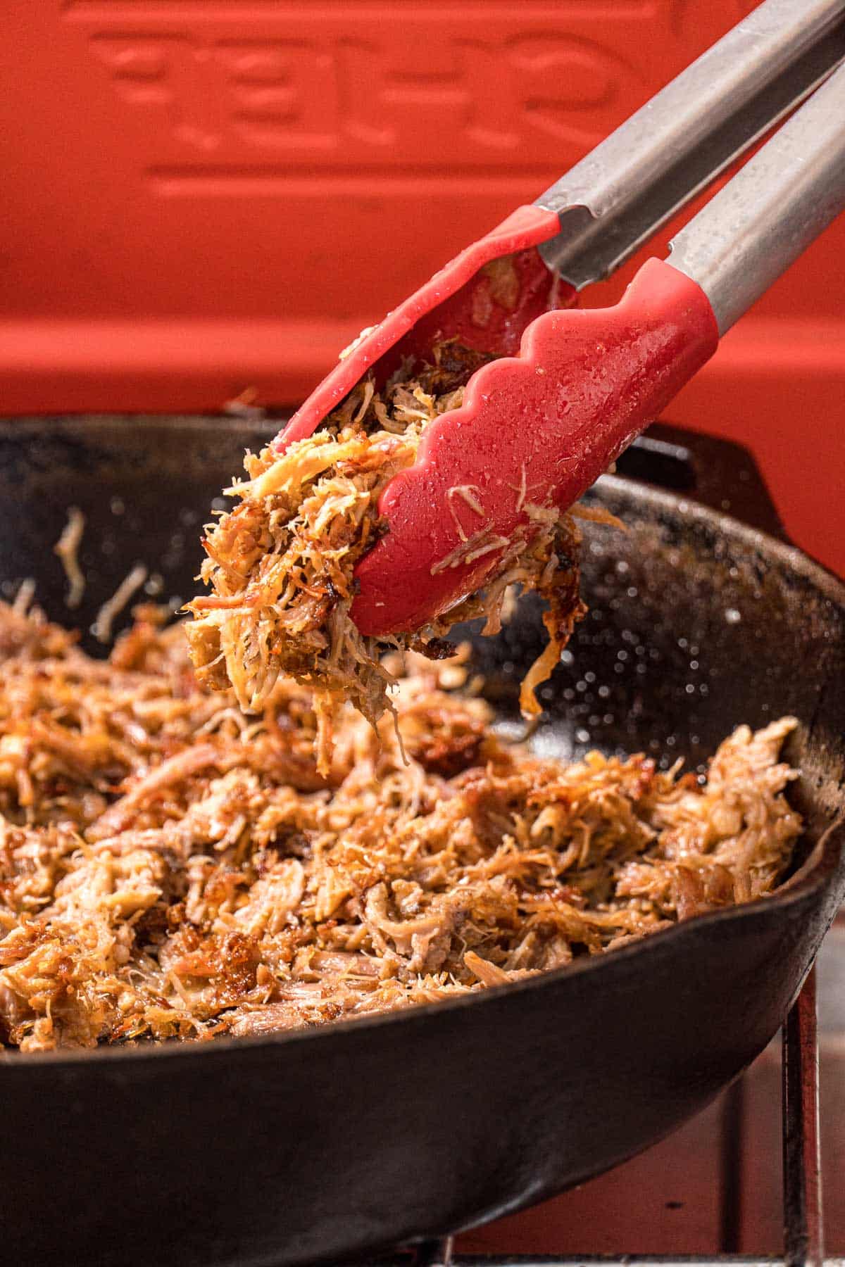 A pair of tongs lifting crispy carnitas out of a cast iron skillet.