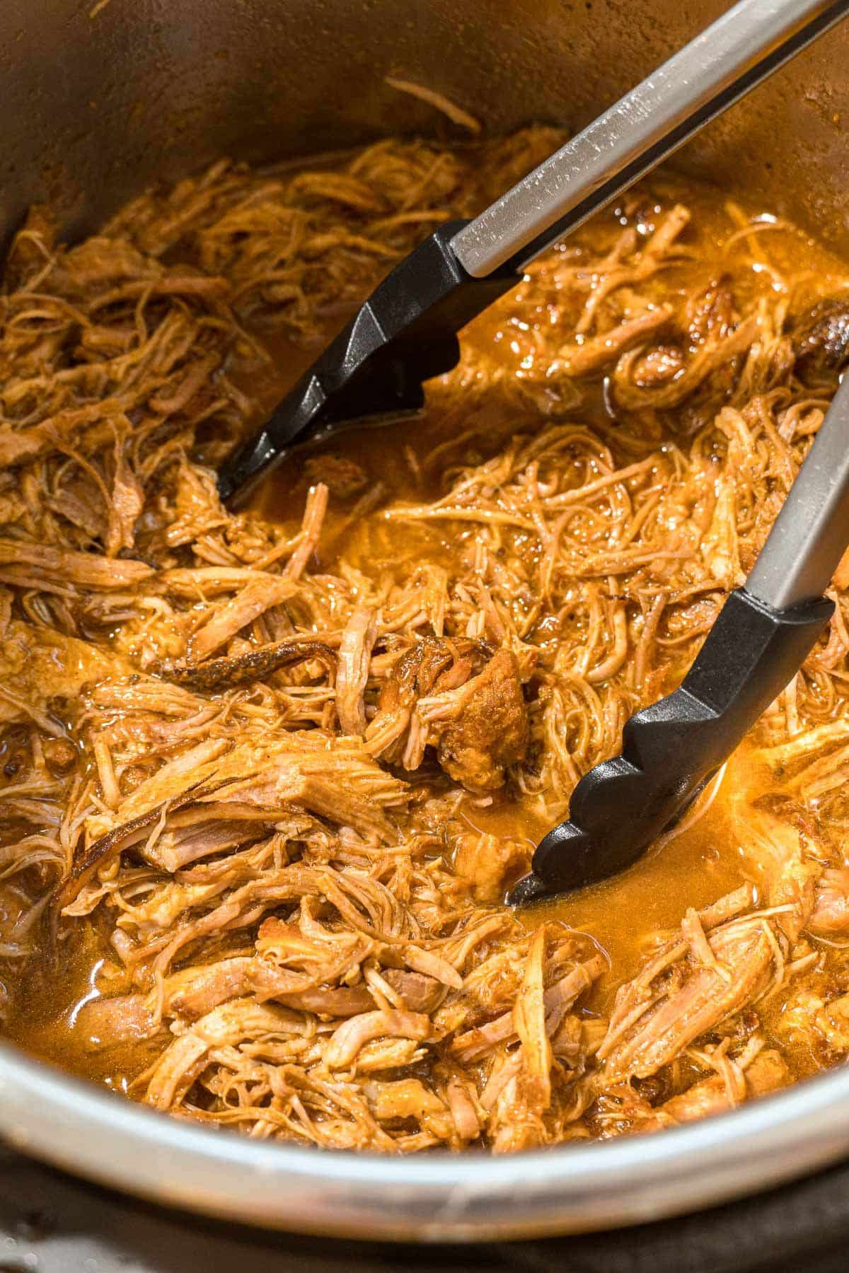BBQ pulled pork in an Instant Pot with a pair of tongs.