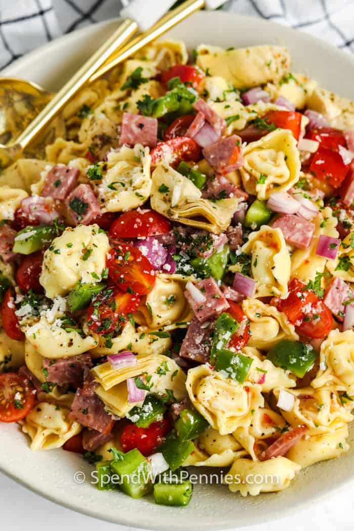 A vibrant plate showcases tortellini, cherry tomatoes, diced ham, and artichokes sprinkled with chopped parsley and dusted with grated cheese, accompanied by gold forks.