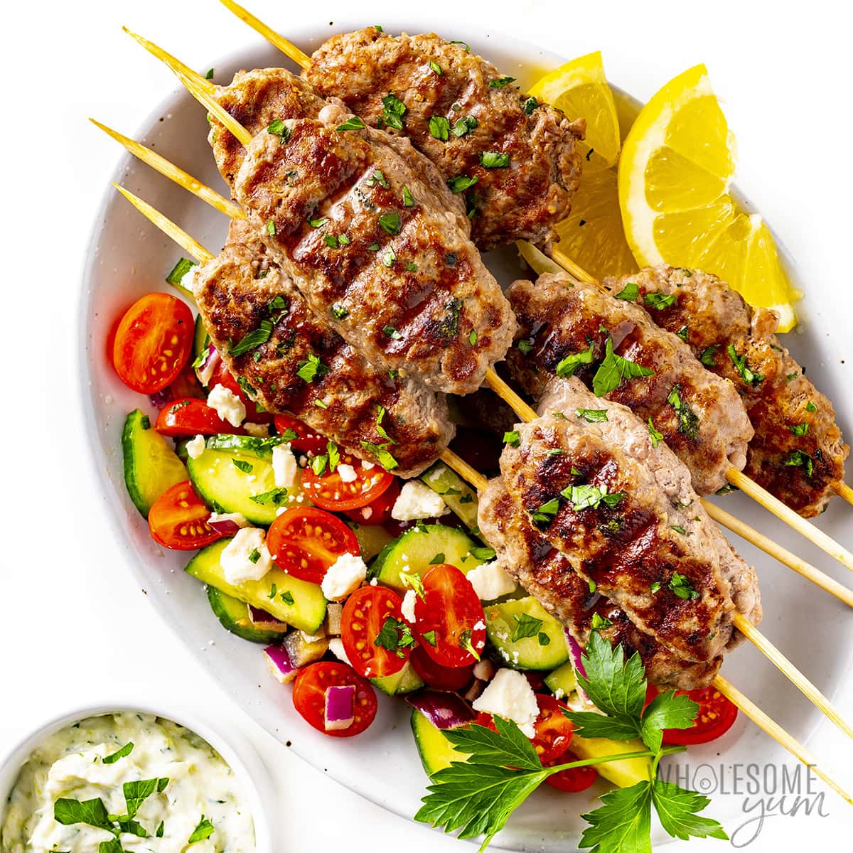 Grilled kebabs, cucumber tomato salad, and tzatziki sauce are presented on a white platter, accented with lemon wedges and herbs.