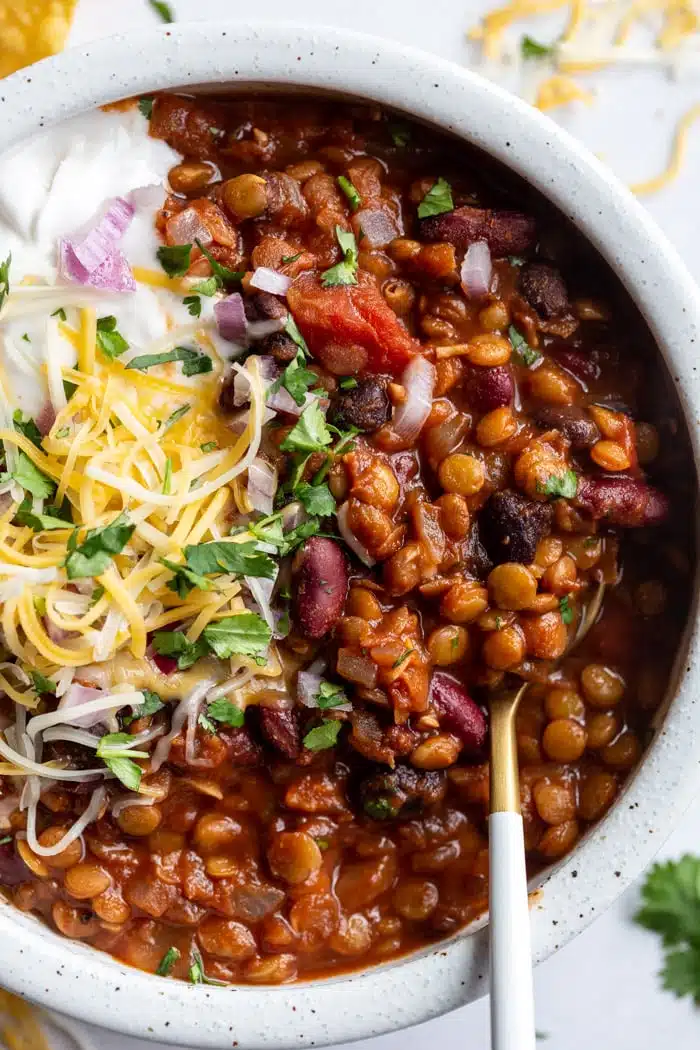 A bowl is filled with chili, topped by shredded cheese, a scoop of sour cream, diced red onions, and fresh cilantro; a golden spoon rests beside it.