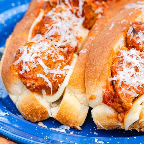 Two meatball subs with tomato sauce and shredded cheese in toasted buns sit on a blue plate, with melted cheese under the meatballs.