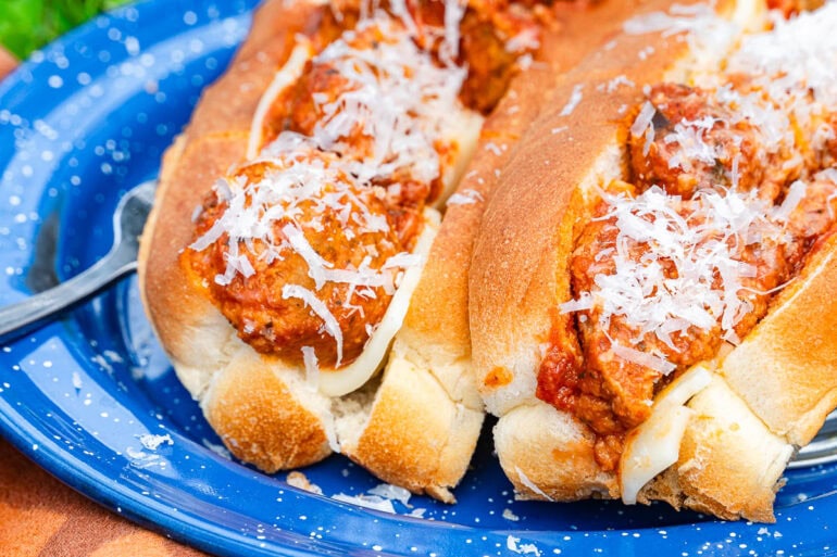 Two meatball subs with tomato sauce and shredded cheese in toasted buns sit on a blue plate, with melted cheese under the meatballs.