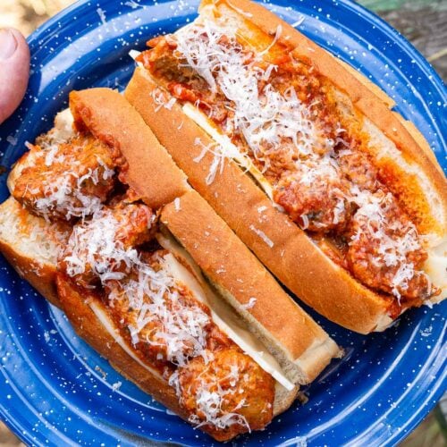 Two hoagie rolls with meatballs, sauce, and grated cheese sit on a blue plate atop a wooden outdoor table.