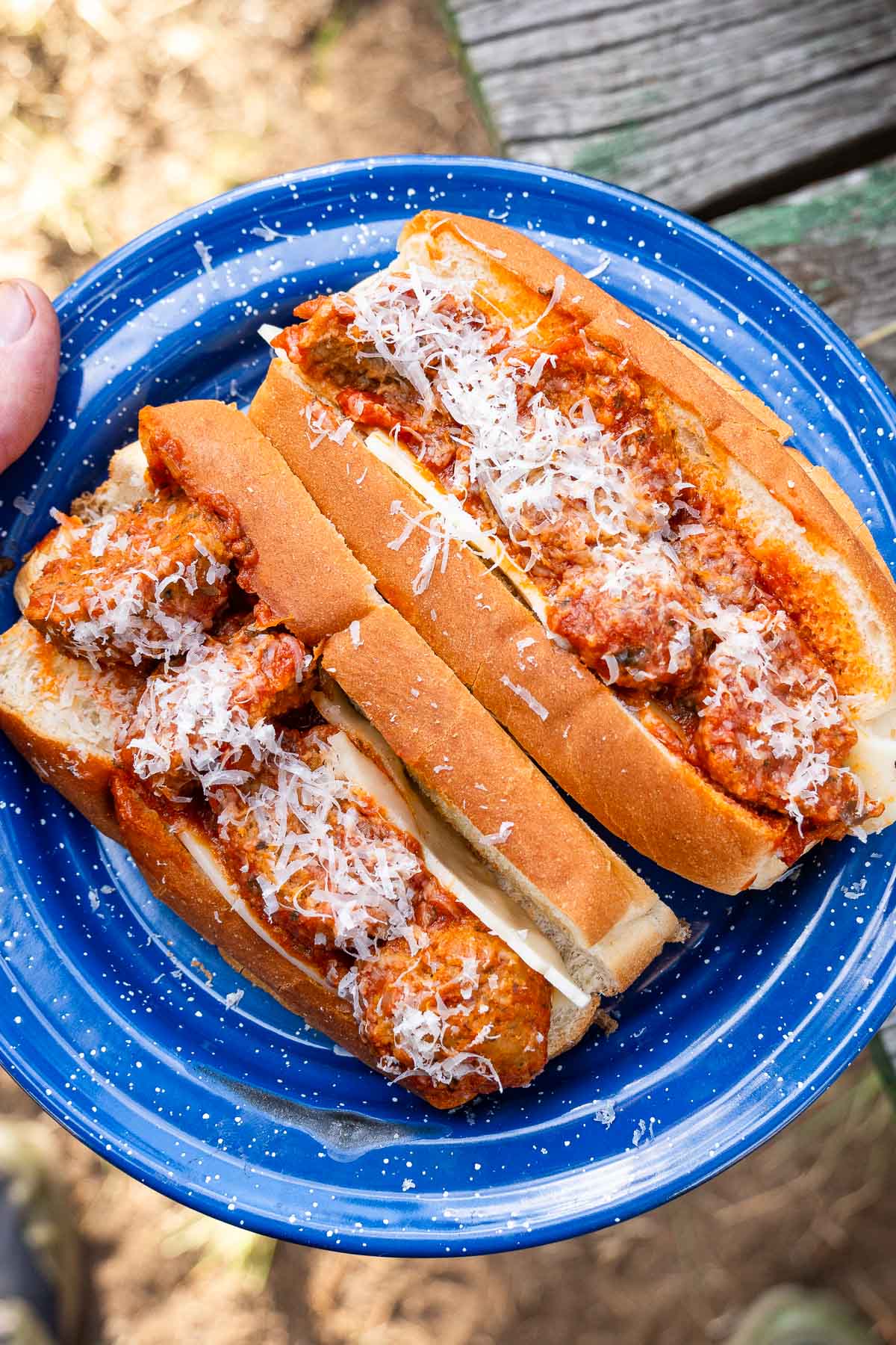 Two hoagie rolls with meatballs, sauce, and grated cheese sit on a blue plate atop a wooden outdoor table.