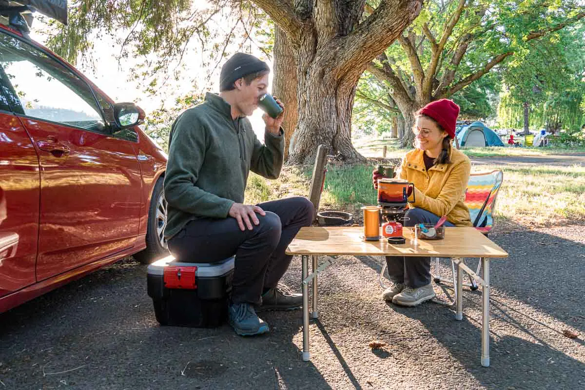 Megan and Michael sit at a small camping table and are drinking cups of coffee.