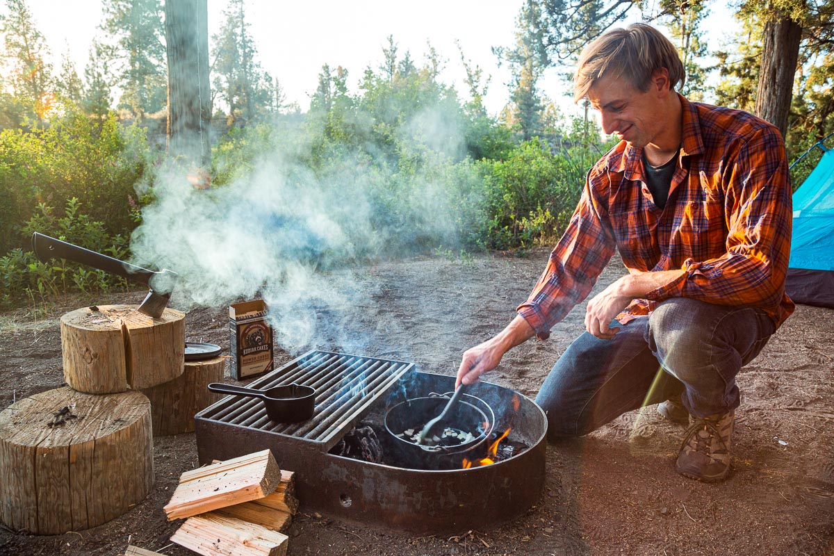 Michael crouched next to a campfire stirring food in a Dutch oven