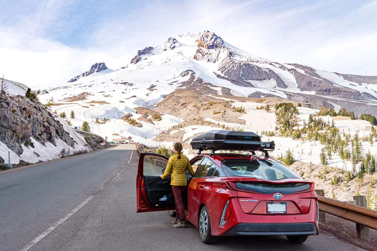A red car is parked on the site of a road. Megan is stepping out of the car to look at Mt. Hood.