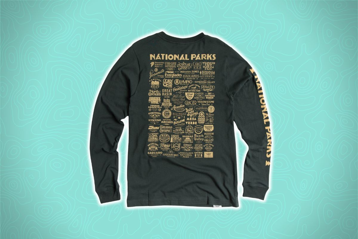 A long-sleeve black shirt with yellow "NATIONAL PARKS" text and icons on the back and sleeve, on a teal topographic background.
