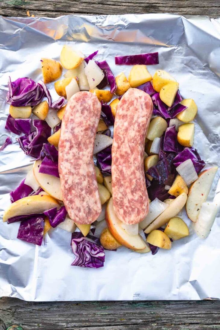 Two raw sausages on aluminum foil, surrounded by chopped purple cabbage, yellow potatoes, apples, and onions on a wooden surface.
