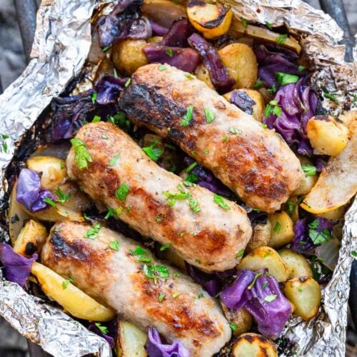 Three grilled sausages on roasted potatoes, purple cabbage, and onions in a foil packet with herbs, set on a barbecue grill.