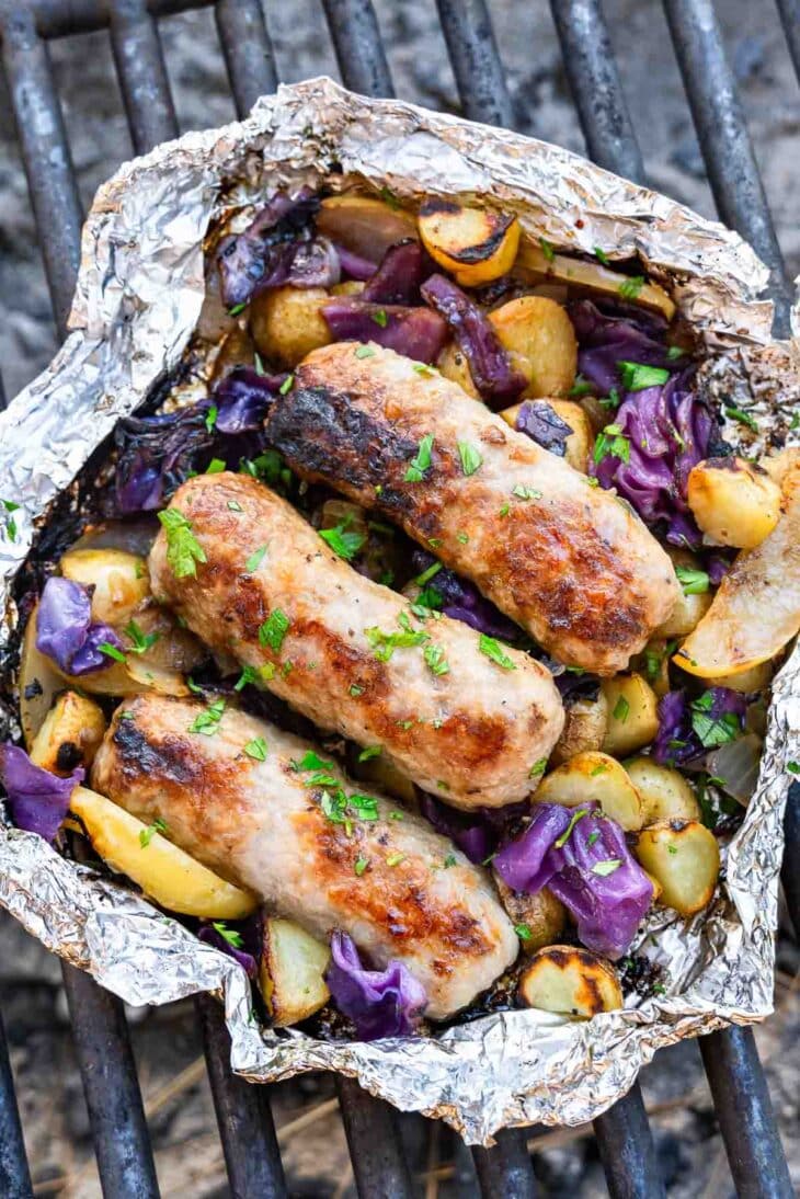 Three grilled sausages on roasted potatoes, purple cabbage, and onions in a foil packet with herbs, set on a barbecue grill.