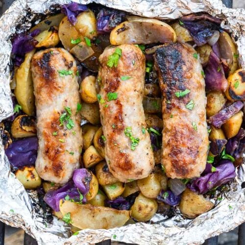 Three grilled sausages rest on roasted potatoes, onions, and purple cabbage in a foil packet on a grill, topped with chopped herbs.