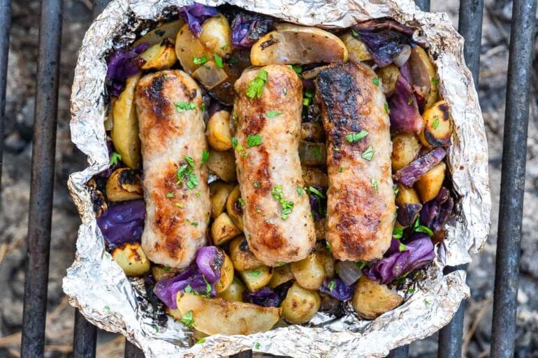 Three grilled sausages rest on roasted potatoes, onions, and purple cabbage in a foil packet on a grill, topped with chopped herbs.
