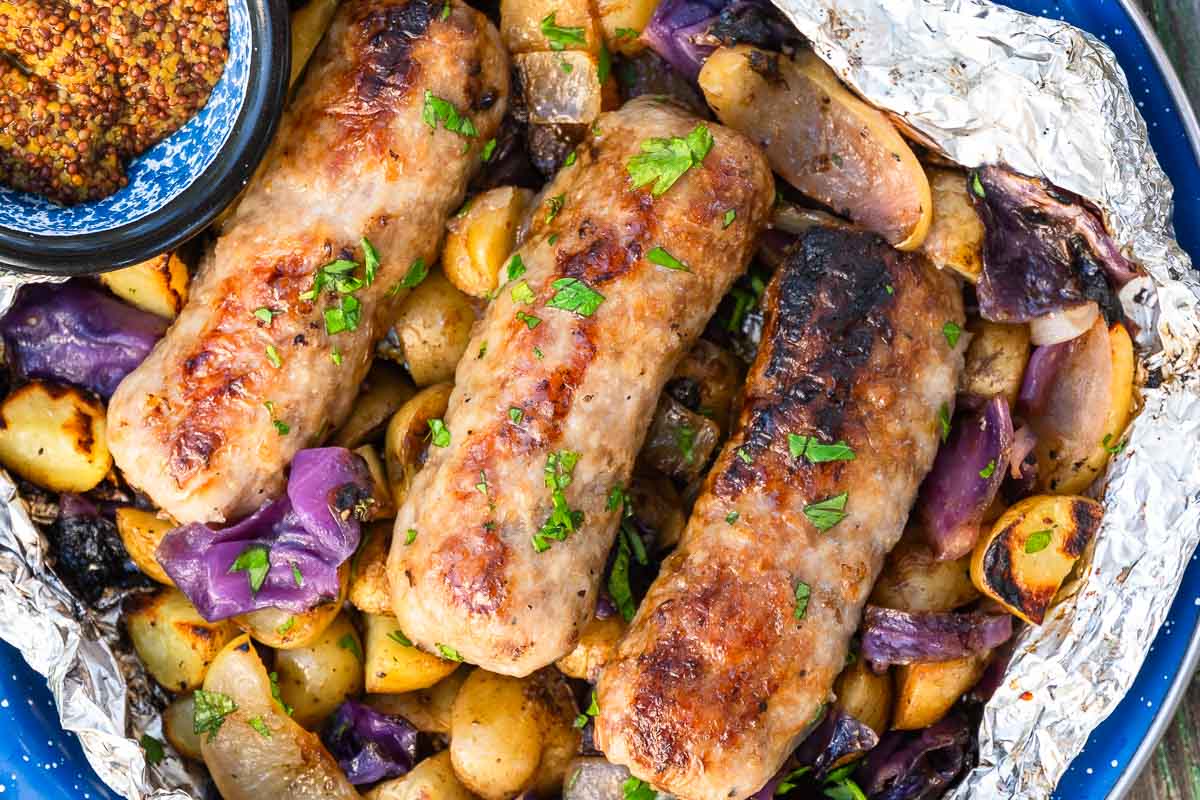 Three browned sausages on roasted potatoes, purple cabbage, and onions in foil with parsley, plus a small bowl of whole grain mustard.