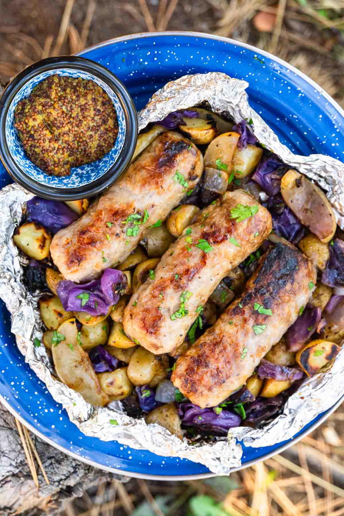 Grilled sausages sit on a foil packet with roasted potatoes, purple cabbage, and herbs on a blue plate beside grainy mustard.