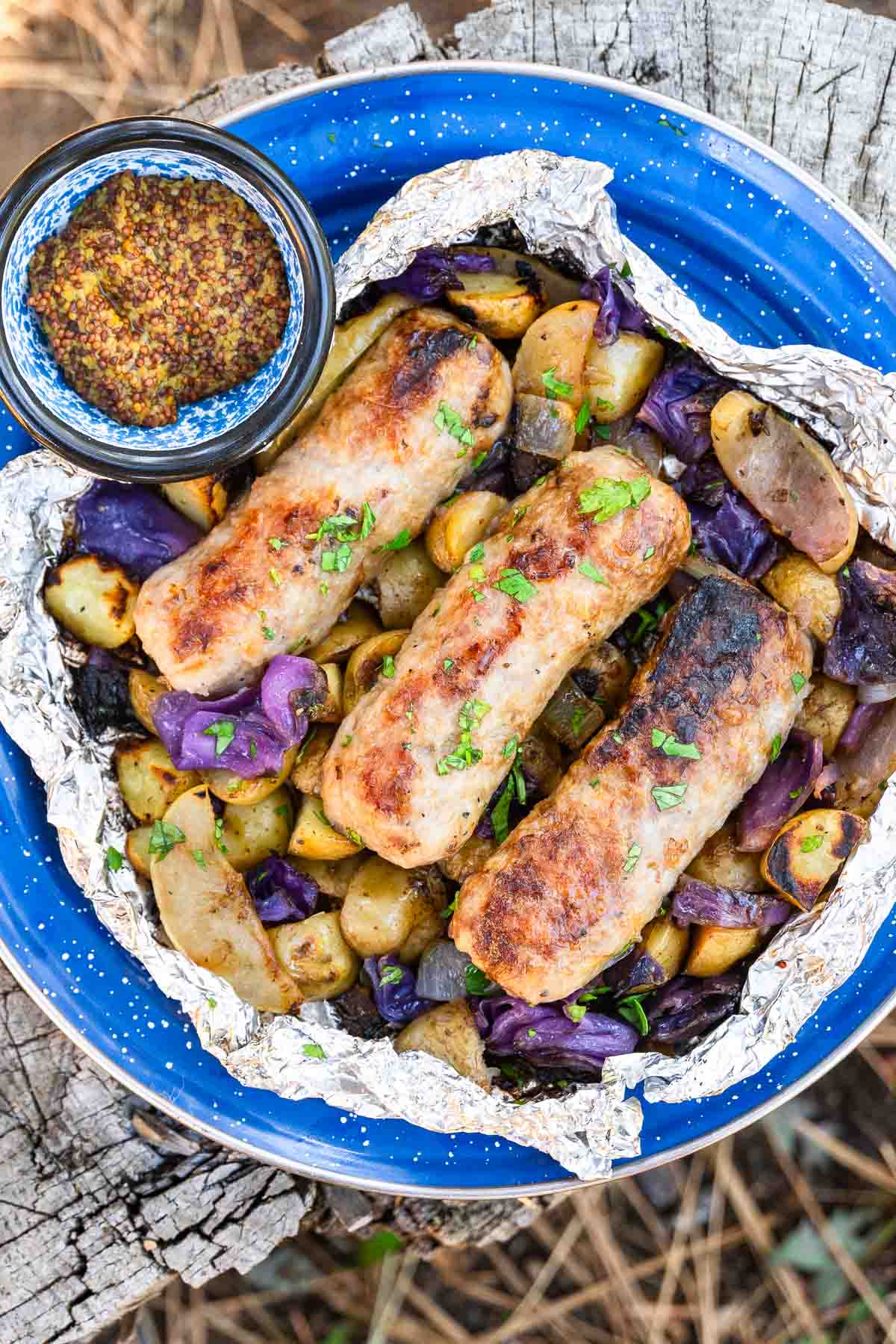 Grilled sausages on roasted potatoes and red cabbage in foil, topped with herbs, plus mustard in a jar on a blue enamel plate outdoors.