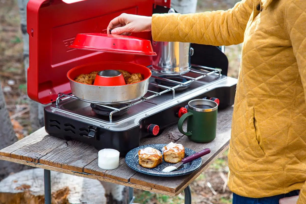An Omnia stovetop oven on a camping stove. Megan is lifting the lid to show the cinnamon rolls inside.