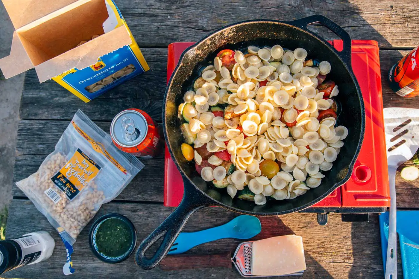 Overhead view of pasta in a cast iron skillet on a camping stove. Other pasta ingredients such as cheese, pesto, and pine nuts are on the table.