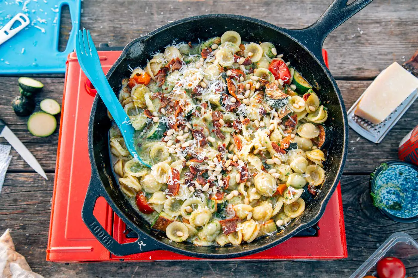 Overhead shot of pesto pasta in a cast iron skillet on a camping stove topped with cheese and pine nuts.